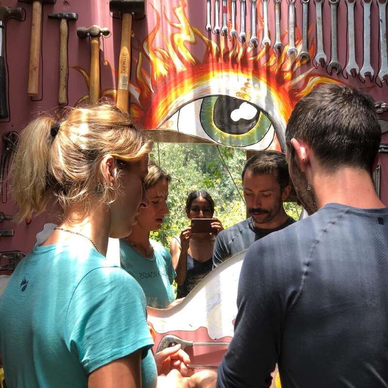 Drei Personen stehen vor einem skurrilen Spiegel, der mit Werkzeugen und einem gemalten feurigen Auge umrahmt ist. Eine Person macht ein Foto, das ihr Spiegelbild und die lebendige, künstlerische Umgebung festhält.