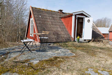 Une petite cabane en bois rouge au toit en pente se dresse dans un cadre rustique. Devant, une table et deux chaises sont posées sur un terrain rocailleux.