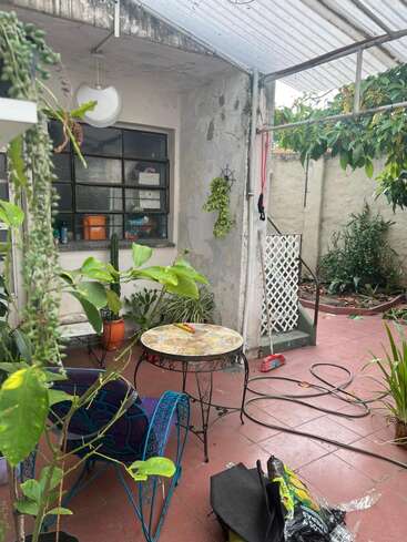 Um pátio aconchegante, cheio de plantas, com uma pequena mesa redonda e uma cadeira azul. Ferramentas de jardinagem, mangueira, vassoura e vários vasos criam uma atmosfera relaxante, meio ao ar livre.