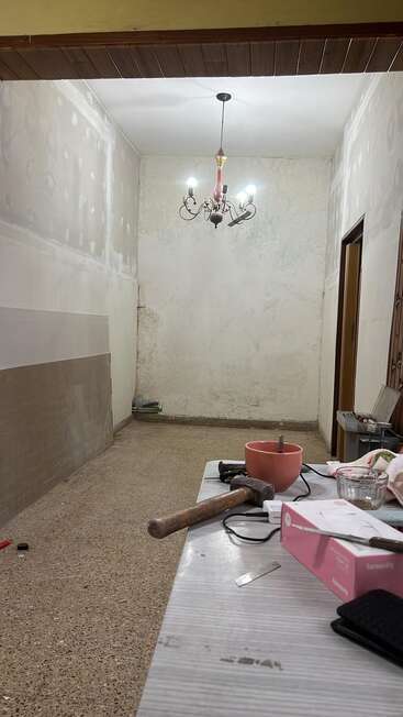A imagem mostra um quarto parcialmente reformado com paredes inacabadas, um lustre, ferramentas e materiais sobre uma mesa, e vários itens espalhados pelo espaço.