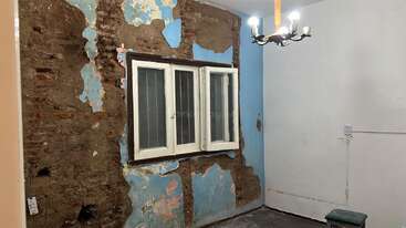 Um pequeno quarto com tinta azul danificada e descascada, tijolos à vista e uma janela com moldura branca. Um lustre está pendurado no teto e um banco verde fica ao lado.