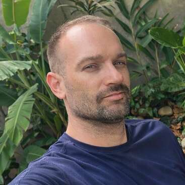 Un homme aux cheveux courts et à la barbe taillée porte une chemise bleu marine. Il pose à l'extérieur devant des plantes vertes feuillues, en regardant l'appareil photo.