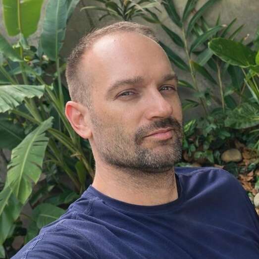 Um cara de cabelo curto e barba aparada está usando uma camisa azul marinho. Ele está posando ao ar livre, na frente de plantas verdes, olhando para a câmera.