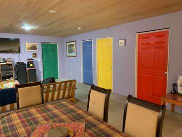The image depicts a dining room with a table, chairs, and colorful doors, featuring a TV, speakers, and a couch in the background.