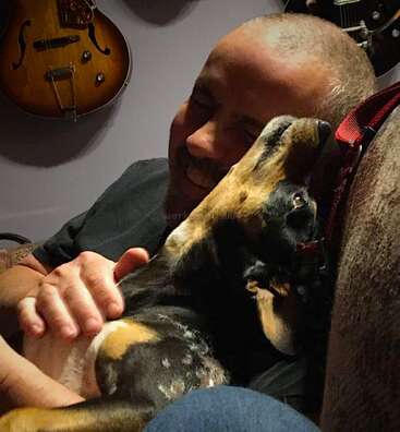 A joyful man cuddles closely with a happy, relaxed dog on a couch. Warm lighting and guitars hanging on the wall create a cozy atmosphere.