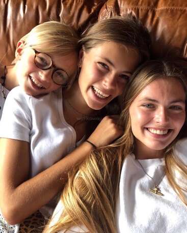 La imagen muestra a tres chicas jóvenes tumbadas en un sofá de cuero marrón, sonriendo a la cámara, con camisas blancas y collares.
