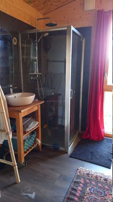 This cozy bathroom features a glass shower, wooden sink stand with towels, large mirror, red curtain, window for natural light, dark flooring, and patterned rug.