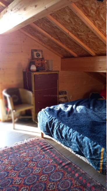 A cozy attic bedroom with wooden walls and slanted ceiling, a patterned rug, blue blanket, chair, wooden dresser, and warm sunlight streaming in.