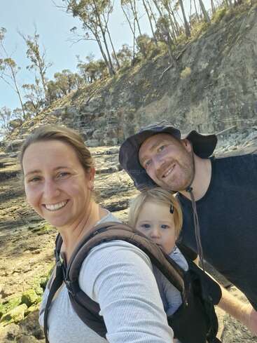 A smiling woman carries a baby in a front carrier, standing beside a man in a sunhat. They are hiking outdoors near rocky cliffs and trees.