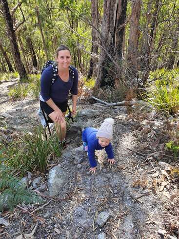 A woman with a backpack smiles at the camera while hiking in a forest. A toddler in a beanie crawls playfully beside her among trees and rocks.