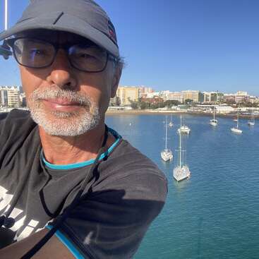 La imagen representa a un hombre con sombrero y gafas, con una serena masa de agua con varios barcos al fondo, sobre un cielo azul despejado.