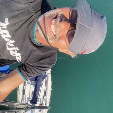 La imagen muestra a un hombre con sombrero y gafas de sol, de pie en un barco con un velero blanco de fondo, sobre una masa de agua azul verdosa.