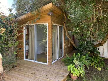 Una acogedora cabaña de madera con grandes puertas correderas de cristal se encuentra entre frondosos árboles, rodeada de una pequeña terraza y macetas.