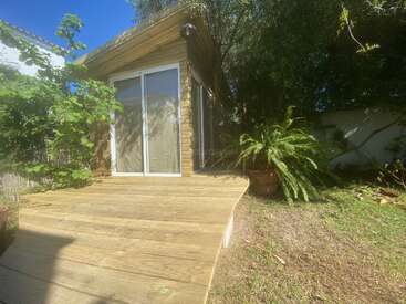 Una pequeña cabaña de madera con puertas correderas de cristal se asienta sobre una soleada terraza de madera, rodeada de frondosas plantas y árboles, creando un acogedor refugio en el jardín.