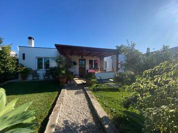 Una encantadora casa blanca con un porche de madera, rodeada de exuberante vegetación y plantas, un cielo soleado en lo alto, acogedoras sillas y un camino de grava que atraviesa el jardín.