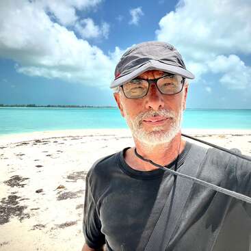 Un hombre con gafas y gorra gris está de pie en una playa soleada con agua turquesa, arena blanca y un cielo azul parcialmente nublado de fondo.