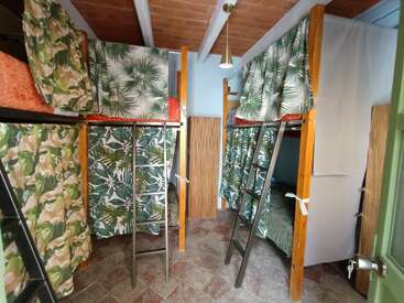 This image shows a bright hostel dorm room with wooden bunk beds, leafy-patterned privacy curtains, metal ladders, tiled floor, and a light fixture hanging from the ceiling.