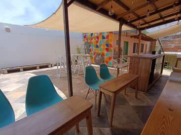 This image shows a modern outdoor patio with blue chairs, wooden tables, a bar, colorful mural, shade sails, and a bright, inviting atmosphere.