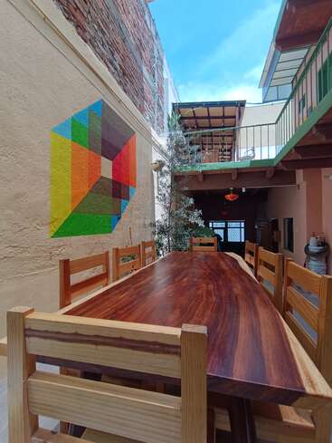 A long wooden table with matching chairs sits in a cozy courtyard, featuring a colorful geometric mural on a brick wall under a bright blue sky.