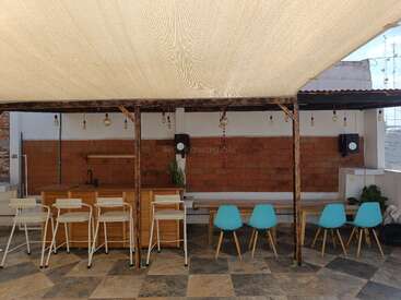 This image shows a shaded outdoor seating area with a bar and kitchen counter, white and blue chairs, hanging light bulbs, speakers, and tiled floor.
