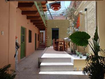This image shows a sunny, cozy courtyard with plants, wooden stairs, green doors, a dining table, warm-colored walls, and a modern hanging lamp overhead.