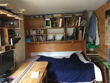 In diesem gemütlichen, kompakten Zimmer stehen ein Holzbett mit einer blauen Decke, ein unordentlicher Schreibtisch, Bücherregale voller Bücher, eine Lampe und hängende Kleidung.
