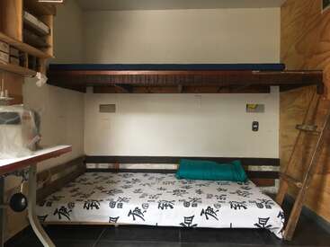 This image shows a small room with a wooden bunk bed setup, a ladder, patterned bedspread, a turquoise pillow, shelves, and a sewing machine workspace.