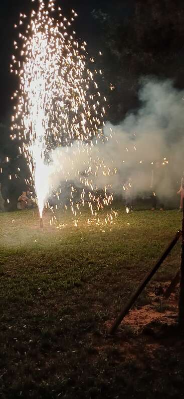 Ein leuchtendes Feuerwerk schießt Funken und Rauch in den Nachthimmel über einer grünen Wiese, während im Hintergrund Menschen zuschauen und draußen feiern.