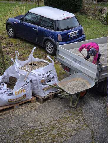 Ein Kind in einer rosa Jacke schaufelt Kies von einem Anhänger in eine Schubkarre. Daneben stehen große Kiessäcke und ein blauer Mini Cooper.