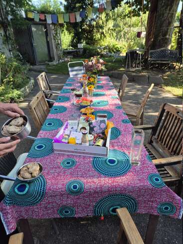 Ein langer, bunter Tisch im Freien ist mit Snacks, Getränken und Blumen gedeckt. Holzstühle stehen drum herum, und über dem Tisch hängen im Garten Fahnenschlangen.