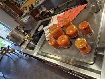Sieben Gläser mit hausgemachter Tomatensauce kühlen kopfüber auf einer Arbeitsplatte aus Edelstahl in einer gemütlichen, rustikalen Küche mit Töpfen, Handtüchern und Holzmöbeln.
