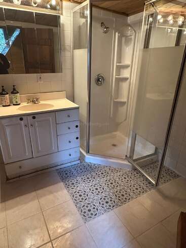 This bathroom features a corner glass shower, white vanity with sink, mirrored medicine cabinet, patterned tile accent floor, and wooden ceiling, creating a cozy, rustic feel.