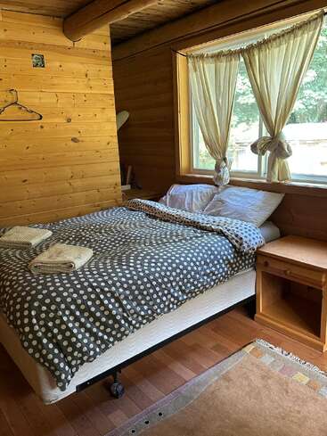 A cozy wooden cabin bedroom with a polka dot comforter, two folded towels, large window with tied curtains, small nightstand, and natural sunlight streaming in.