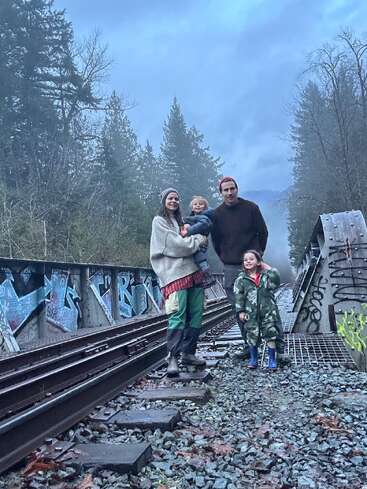 A family of four stands on a graffiti-covered railway bridge surrounded by tall trees and mist. They wear warm clothes and look happy together outdoors.
