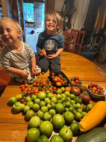 Two smiling children sit on a wooden table surrounded by freshly picked garden vegetables—green tomatoes, zucchini, and colorful cherry tomatoes—creating a joyful, homegrown harvest scene indoors.