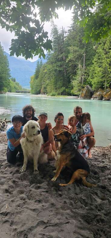 A group of people and two dogs pose on a sandy beach by a beautiful river, surrounded by lush green forest and distant mountains under cloudy skies.