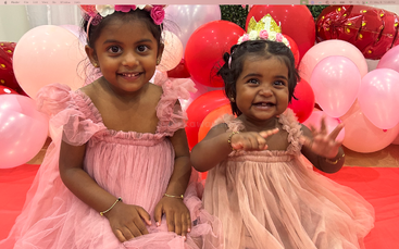 Two adorable young girls in matching pink dresses sit on a red carpet, surrounded by festive red and pink balloons, smiling joyfully at the camera.