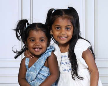 Two young girls with dark hair and ponytails smile brightly. One wears a blue dress, the other a white dress. They have their arms around each other.
