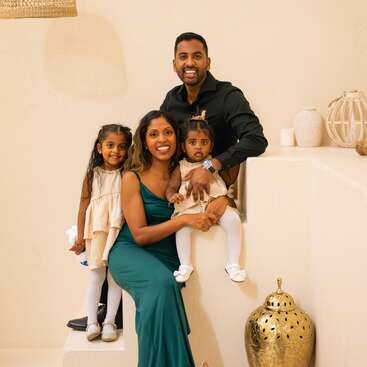 A smiling family of four poses indoors. The father wears black, the mother in green, and their two young daughters wear matching cream dresses and tights.