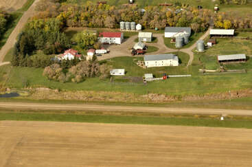 This aerial image shows a rural farmstead with various buildings, silos, barns, trees, machinery, and green fields, situated near a road and farmland.
