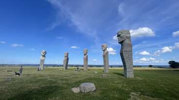 Cinq grandes sculptures en pierre représentant des têtes humaines se tiennent debout dans un champ herbeux sous un ciel bleu lumineux, entourées de rochers, de personnes et d'un rivage lointain.