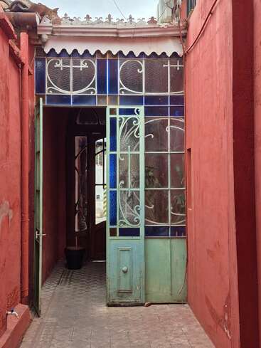 A narrow passage with red walls leads to a vintage, ornate glass door framed by blue and white patterns, opening into a tiled interior space.