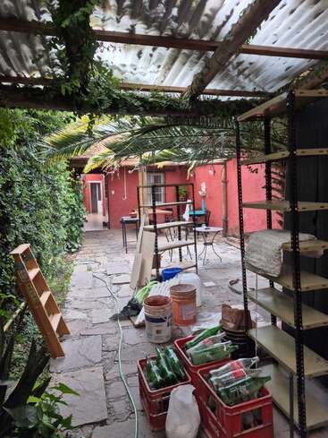A covered patio with greenery, crates of glass bottles, buckets, a ladder, and shelves, indicating ongoing maintenance or renovation. The path leads to a red-walled building.
