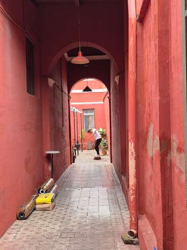 A narrow corridor with red walls and arched ceilings. A person stands in the distance, bending down. Rolled materials and boxes are scattered on the tiled floor.