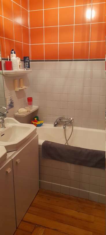 Dieses Badezimmer verfügt über leuchtend orangefarbene Wandfliesen, weiße Armaturen, eine Badewanne mit Handbrause, einen Holzboden, Regale, Pflegeprodukte, ein Handtuch und buntes Badespielzeug.