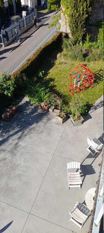 Das Bild zeigt einen sonnigen Innenhof mit weißen Stühlen, Topfpflanzen, einer Rasenfläche, einem roten Klettergerüst, zwei spazierenden Personen und einem Auto in der Nähe.