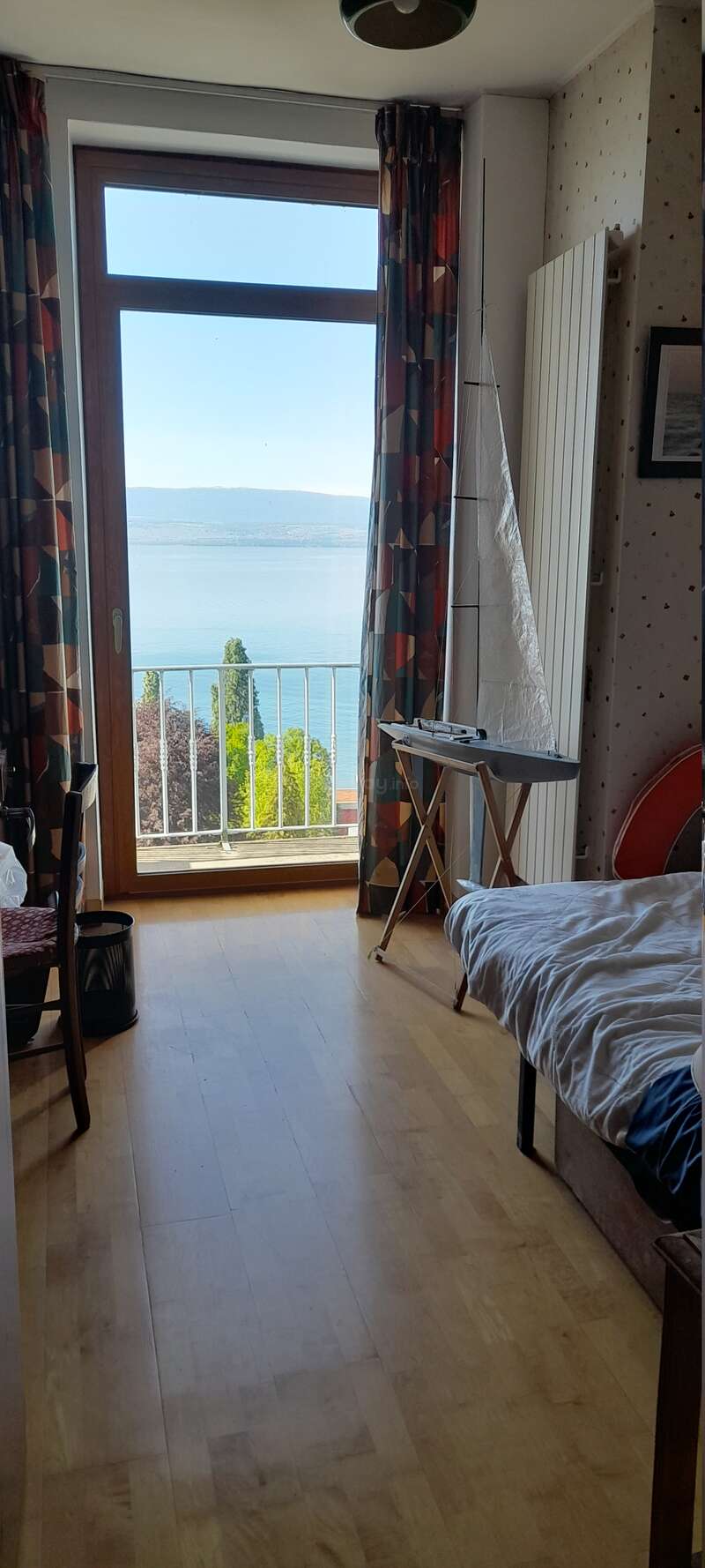 Ein gemütliches Zimmer mit Holzboden, Bett, bunten Vorhängen und einem Segelbootmodell. Durch das große Fenster hat man einen atemberaubenden Blick auf den See und die Berge. Ruhige Atmosphäre.