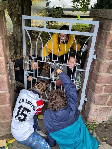 Five people work together, painting or repairing a metal gate fixed between two stone pillars. They are outdoors, wearing casual clothing, focused on the task, collaborating enthusiastically.