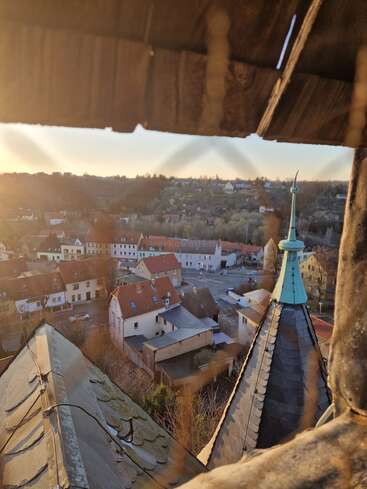 The image shows a scenic view from a high vantage point, overlooking rooftops, a church steeple, and a sunlit town surrounded by hills at sunset.