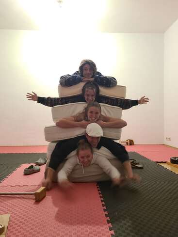 Five people are stacked on top of each other with mattresses in between, smiling and laughing, in a room with foam tiles and casual, playful energy.